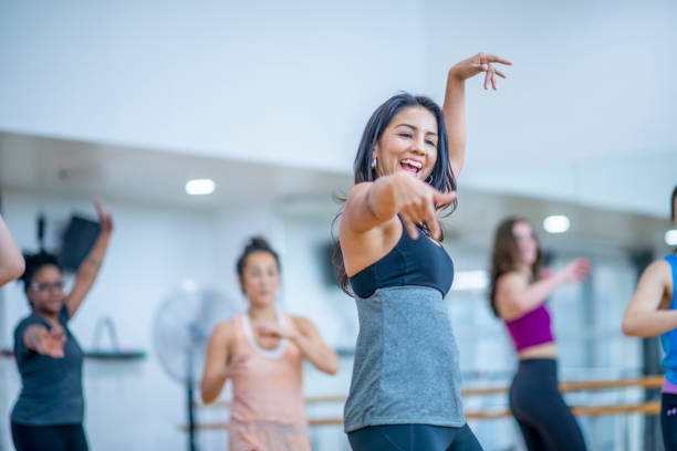 A multi-ethnic group of adult women are dancing in a fitness studio. They are wearing athletic clothes. An Ethnic woman is smiling at the camera while dancing.
