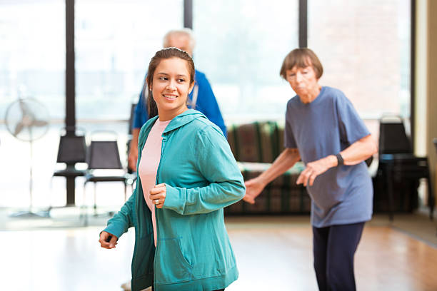 Young Hispanic woman watches the instructor in dance class at local gym. A senior couple is dancing behind her. They are in the exercise studio at the gym. Large bright windows are behind them.