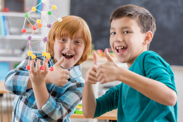 Cheerful elementary school boys give a thumbs up after completing an engineering project at school. One boy is holding the project made from toothpicks and gum drop candy.