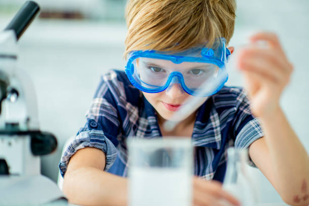 An elementary school is indoors in her house. She is wearing safety goggles, and taking a sample out of a beaker.
