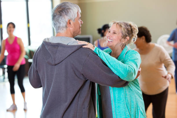 Happy active senior couple take ballroom dancing lessons at senior center. They are in the exercise studio. People are dancing in the background.