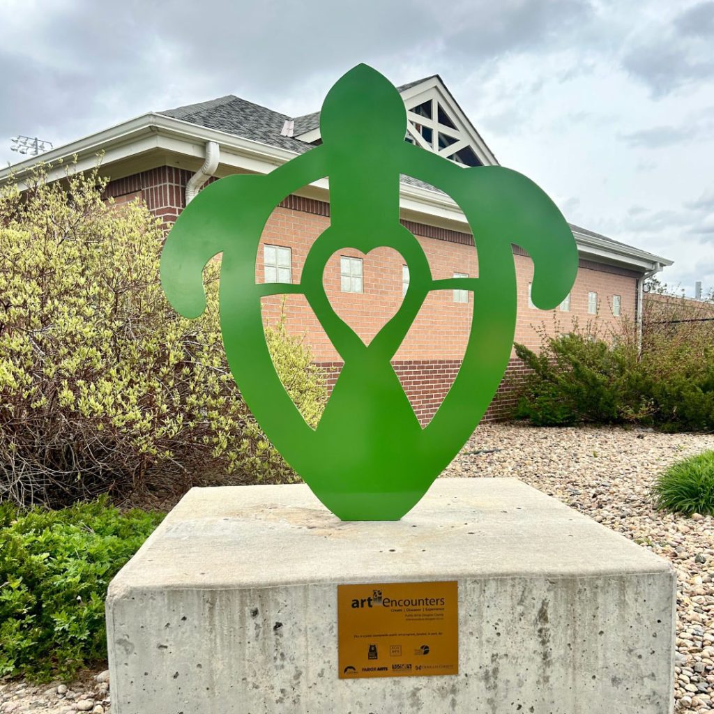 Large outdoor metal 2-D sculpture of a green sea turtle on a concreate pedestal.