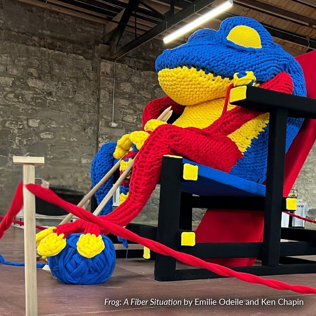 "Frog: A fiber Situation" by Emilie Odeile and Ken Chapin. A big blue, red and yellow frog made of fiber yarn is sitting in a chair, knitting a giant yarn ball of his own.