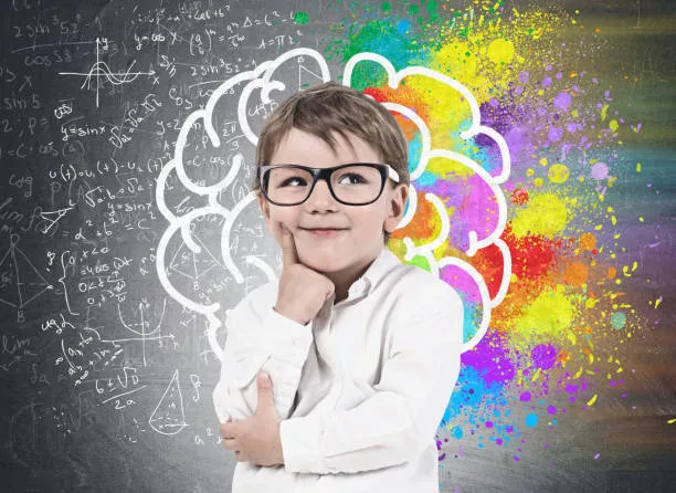 Smiling thinking little boy wearing white shirt and glasses standing near blackboard with colorful brain sketch drawn on it. Concept of creative thinking